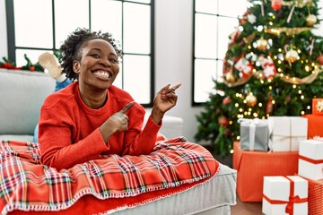 Canvas Print - Young african american woman lying on the sofa by christmas tree smiling and looking at the camera pointing with two hands and fingers to the side.