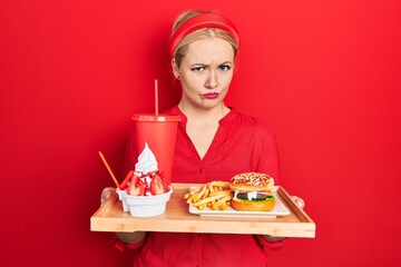 Canvas Print - Young blonde woman eating a tasty classic burger with fries and soda skeptic and nervous, frowning upset because of problem. negative person.