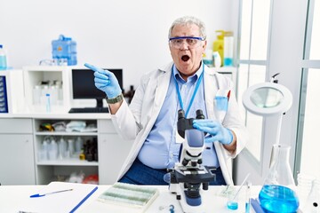 Canvas Print - Senior caucasian man working at scientist laboratory surprised pointing with finger to the side, open mouth amazed expression.