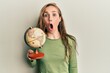 Young blonde woman holding vintage world ball scared and amazed with open mouth for surprise, disbelief face