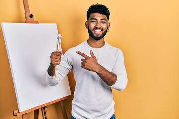 Sticker - Arab man with beard standing by painter easel stand holding brushes smiling happy pointing with hand and finger