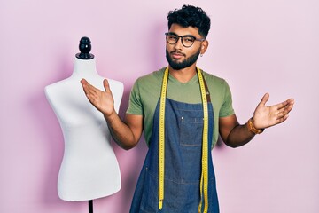 Wall Mural - Arab man with beard dressmaker designer wearing atelier apron clueless and confused with open arms, no idea and doubtful face.