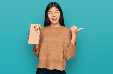 Canvas Print - Beautiful young asian woman holding gift pointing thumb up to the side smiling happy with open mouth