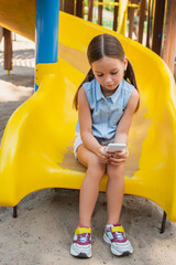 Wall Mural - full length view of girl in summer clothes playing on smartphone while sitting on playground.