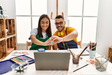 Canvas Print - Young hispanic couple using laptop sitting on the table at art studio smiling in love showing heart symbol and shape with hands. romantic concept.