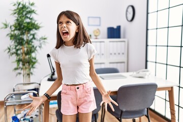 Wall Mural - Young hispanic girl standing at pediatrician clinic angry and mad screaming frustrated and furious, shouting with anger. rage and aggressive concept.