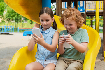 Wall Mural - excited brother and sister sitting on slide and playing on smartphone.