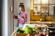 © rh2010 - Woman using digital tablet hanging on the wall in the kitchen, controlling smart home devices. Modern kitchen interior with food in front