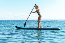Young female surfer in bikini riding standup paddleboard in ocean.
