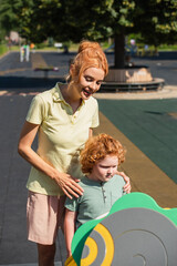 Wall Mural - happy redhead woman hugging shoulders of son on playground.