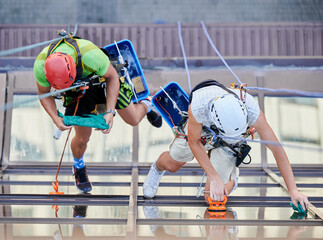 Wall Mural - Industrial mountaineering. Top view of professional cleaners hanging on ropes while cleaning skyscraper windows. Two men window washers using cleaning tools while washing building facade.