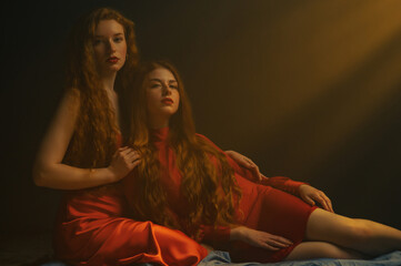 Two young beautiful redhead freckled women with long natural hair wearing red dresses posing in darkness and warm light. Studio art portrait. Copy, empty space for text