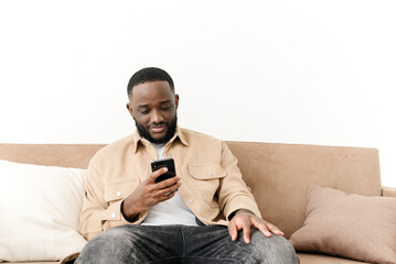 Happy bearded afro american freelancer working using phone while sitting on sofa at his home. Concept of young people working on mobile devices