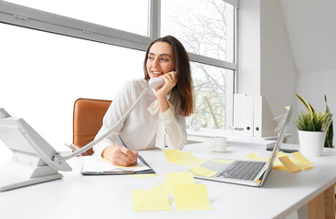 Sticker - Young manager writing on sticky note paper while talking by phone at her workplace