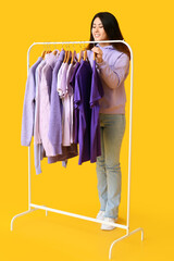 Sticker - Young Asian woman and rack with clothes in purple shades on yellow background