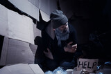 Fototapeta Młodzieżowe - A homeless bearded man sits on boxes on the street and asks for help. Need a homeless person asks for money for food and overnight.