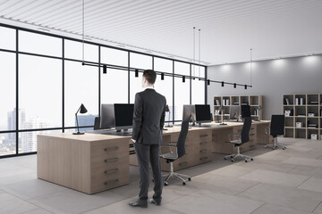 Wall Mural - Young european businessman standing in modern wooden and concrete coworking office interior with furniture and window with city view. Workplace concept.