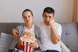 Portrait of bored sad upset wife and husband sitting on sofa with pop-corn and remote control, watching boring movie, looking at camera with frowning faces.