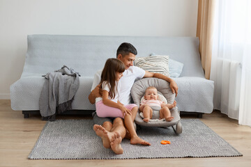 Portrait of brunette man wearing white casual style t shirt sitting on floor near sofa with his daughters, elder child sitting on father legs and looking with dad after sister in rocking chair.