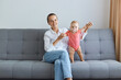 © sementsova321 - Image of positive optimistic woman wearing white shirt and jeans sitting on sofa with her tiny daughter, baby in pink bodysuit standing with mother's help.