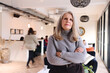 © Southworks - Portrait of confident senior white businesswoman with long gray hair looking at camera with arms folded in coworking space