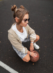 Wall Mural - Fashionable beautiful young hipster girl with vintage sunglasses in stylish clothes with a windbreaker, T-shirt, leggings and sneakers with a ball on the basketball court