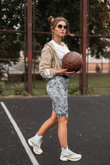 Wall Mural - Fashionable beautiful sportswoman in sportswear with sneakers and a ball playing basketball in the street