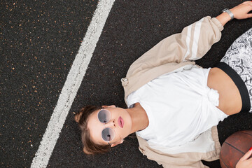 Wall Mural - Fashionable beautiful young woman with retro sunglasses in fashion sportswear with a windbreaker, T-shirt, leggings with a ball lying on the ground on the basketball court, top view