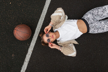 Beautiful hipster woman in fashion sports clothes wearing sunglasses, lying and relaxing on the asphalt of the basketball court, view from above