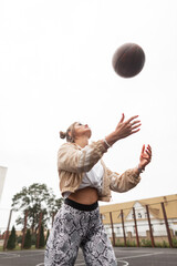 Wall Mural - Beautiful athletic young girl in sportswear with a windbreaker, white t-shirt and leggings plays basketball and throws up the ball on the street