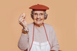 Happy smiling elderly grandmother cook holding an egg, looking at camera. Grey-haired old cook wearing apron and hat at studio over beige background. Copy space.