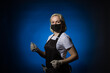 © Sergey - A female gardener in a black apron, gloves in a protective mask walks and looks at the camera on a dark background.