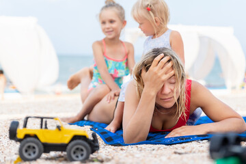 Two cute little sibling girls enjoy having fun playing sitting on tired exhausted mother's back at sea ocean beach. Frustrated mom make face palm gesture. Vacation family small kids trouble concept