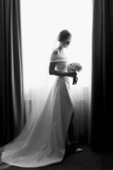  A beautiful bride in a white dress stands near the window and looks at the bouquet. Black and white photo