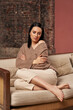 © Elena - a beautiful pensive woman with dark hair sat comfortably on the sofa with her legs crossed.