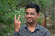 A handsome Indian young man wearing glasses and a shirt showing two fingers or a victory sign and smiling in the garden background