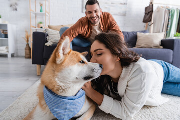 Wall Mural - happy young woman cuddling akita inu dog near blurred boyfriend on couch.