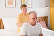 © ulza - elderly woman gives a shoulder massage to her husband sitting on a bed at home. elderly couple healthy lifestyle.