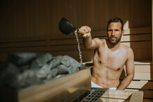 Young Man Pouring Water Onto Hot Stone In The Sauna