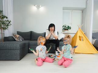 an excited mother in stress looks at two twin girls who are smeared with paint for drawing.