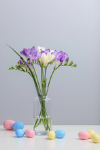 A Bouquet Of Twigs Of Delicate Purple And White Freesia Flower In A Glass Bottle And Colorful Easter Eggs On A Gray Background