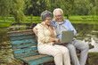 © Studio Romantic - Happy elderly retired husband and wife spend time together, sitting on a bench in the park and looking at the laptop screen, communicating with loved ones at a distance. Outdoor recreation.