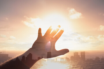 Wall Mural - Hand reaching out to touch the warm rays of sunshine on city sunrise background. Hope, and light concept 