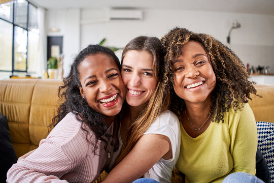Three mixed race happy female friends hugging smiling - Funny women together celebrating sitting on the living room sofa looking at the camera selfie.