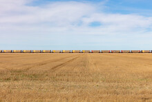 View Across A Stubble Field And The Long Line Of Yellow Boxcar Wagons Of A Freight Train On The Horizon Line. 