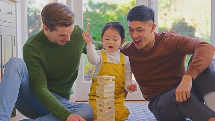 Wall Mural - Family with two dads playing game with daughter at home stacking wooden bricks into tower which fall over- shot in slow motion