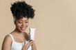 © etonastenka - Bottle of cosmetic cream with a mockup in the hands of an african american female model on a beige background. Lovely black woman advertising moisturizer