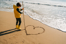 Boy In Yellow Rubber Boots Drawing Heart Shape On Sand At The Beach. School Kid Touching Water At Autumn Winter Sea. Child Having Fun With Waves At The Shore. Spring Holiday Vacation Concept.