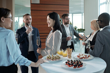 Wall Mural - Three cheerful intercultural colleagues with champagne having discussion of best reports in buffet after conference