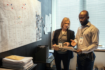 Poster - Two confident agents of FBI in formalwear discussing criminal profiles of suspects while standing in front of board with map
